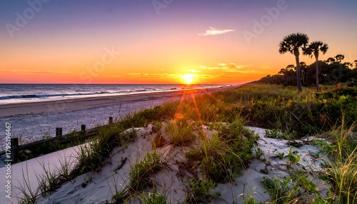 Fototapeta Naklejka Na Ścianę i Meble -  Sunset on a sandy beach with dune grass and palm trees, orange sky, calm water, and wooden barriers