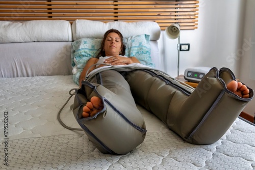 Young woman lying on a bed while undergoing leg compression therapy using a professional pneumatic device at home. Concept of wellness, physiotherapy, lymphatic drainage, muscle recovery