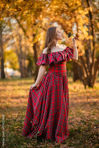 A young woman stands in an autumn forest. She is wearing a long, red, plaid dress with off-the-shoulder details and a frill, a romantic, vintage style. She holds a decorative metal flower in her hands