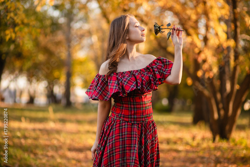 A young woman stands in an autumn forest. She is wearing a long, red, plaid dress with off-the-shoulder details and a frill, a romantic, vintage style. She holds a decorative metal flower in her hands