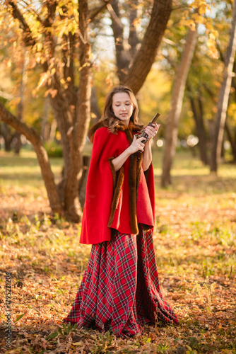 A young woman stands in an autumn forest. She's wearing a long red plaid dress and a warm cape, a romantic, vintage style. She's holding a pistol.