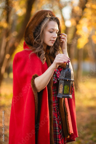 A young woman stands in an autumn forest. She's wearing a long red plaid dress and a warm cape, a romantic, vintage style. She's holding a lantern.