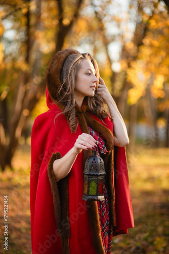 A young woman stands in an autumn forest. She's wearing a long red plaid dress and a warm cape, a romantic, vintage style. She's holding a lantern.