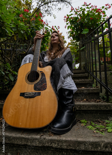 Young woman musician sitting on outdoor steps holding acoustic guitar wearing casual outfit