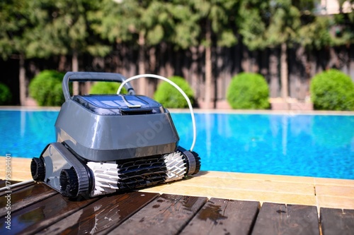 A pool cleaning robot is positioned on the edge of a wooden deck next to the crystal clear blue water. Green shrubs and trees in the background create a cozy, private atmosphere
