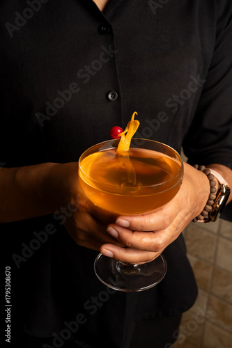 Close-up shot of an orange coloured cocktail in a coupe glass, held in a woman’s hands, featuring an orange peel garnish and a warm, elegant presentation for bar and menu use.