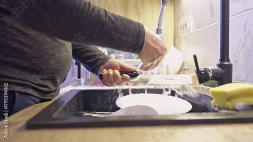 A person diligently washes and rinses dishes under running water in a contemporary kitchen sink, highlighting the routine of domestic cleanliness and home maintenance.