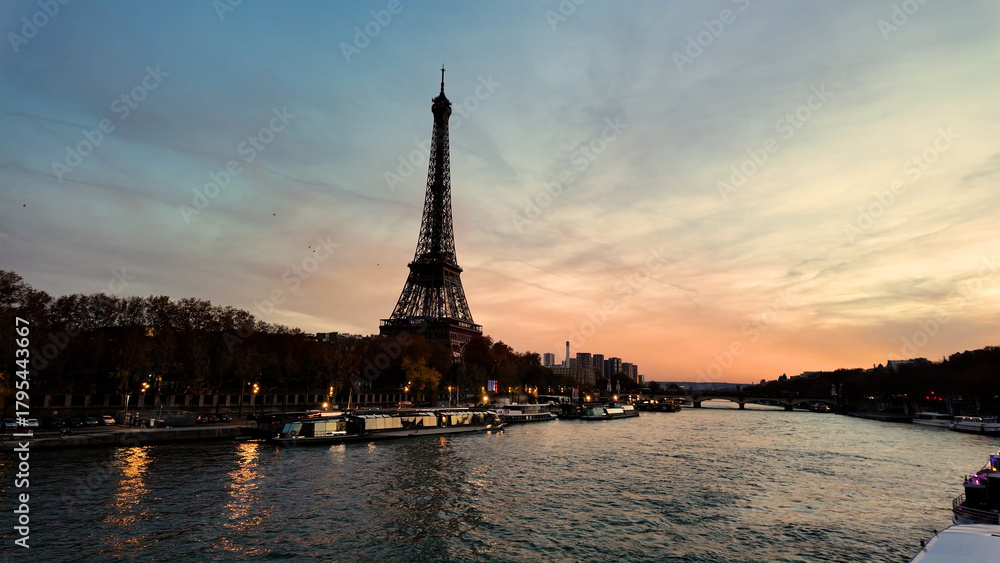 Fototapeta premium A dramatic, high-contrast twilight view of the Eiffel Tower silhouetted against a blue-orange gradient sky, emphasizing the shimmering water reflections of the barges on the Seine.
