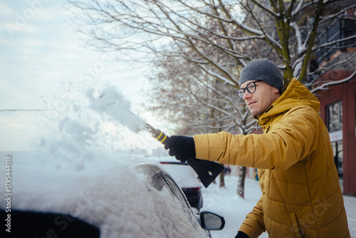 Man in winter clothing clearing snow from car parked in residential city street during cold morning. Scene showing everyday winter routine, weather conditions and urban lifestyle.