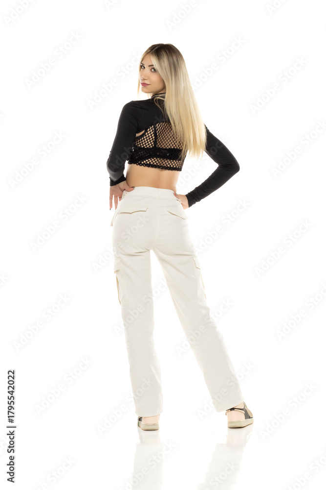 Fototapeta premium Rear view of blonde woman in black long sleeve crop top and white cargo pants looking over shoulder, full body, studio shot on a white background
