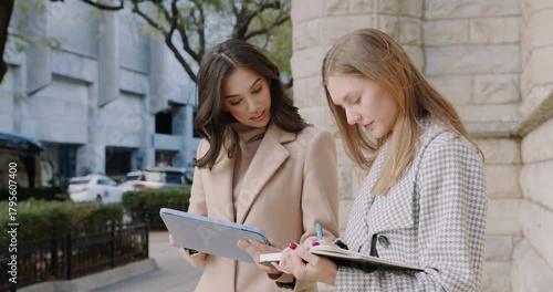 Two professionals review documents in urban setting, Pair of businesswomen collaborating on project near city building