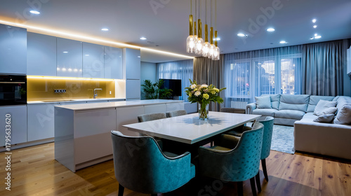A modern kitchen and living room with a white color scheme featuring a dining table with blue chairs a white couch and a chandelier hanging from the ceiling