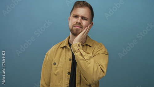 Young man with hand on cheek showing toothache and slight grimace in blue studio portrait; toothache concern discomfort recovery.
