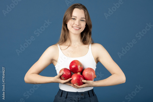 Skincare and healthcare concept. Beautiful young smiling woman with white shirt and blue pants holding red pomegranate fruits a in her hands. Blue background with copy space