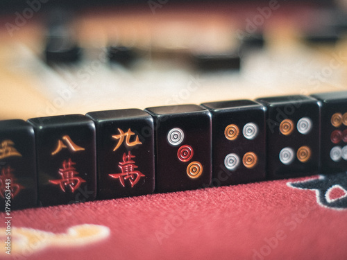 A detailed macro view of black Mahjong tiles arranged in a row on a red felt table, featuring Chinese characters and circular symbols in various colors.