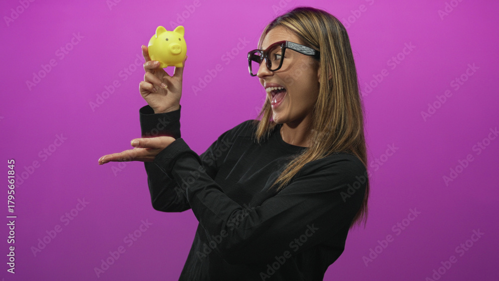 Obraz premium Woman holds yellow piggybank with raised hand and smiles in studio against magenta backdrop; savings confidence.