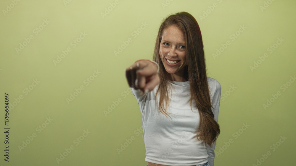 Fototapeta premium Woman pointing finger at camera in studio against lime green wall, bare finger extended and smiling; friendly confidence.