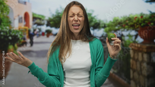 Woman holding lime near face and grimacing with closed eyes and open mouth on street, wearing green cardigan and white shirt; disgust.