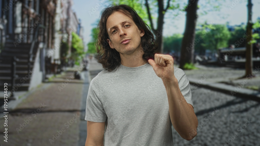 Fototapeta premium Young hispanic man with long hair raising right fist and looking at camera in a city street; confidence defiance determination.