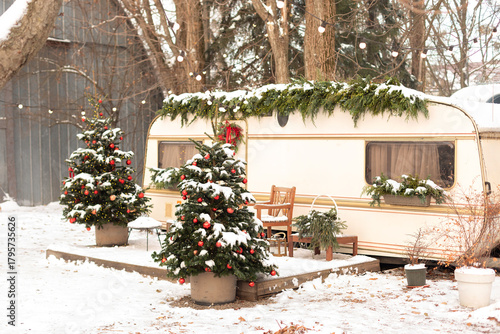 Christmas-themed winter campsite with a festive RV trailer, outdoor firepit, armchair, snow-covered yard, Xmas holiday tree and cozy garden furniture. Exterior Seasonal mobile home getaway. New Year	
