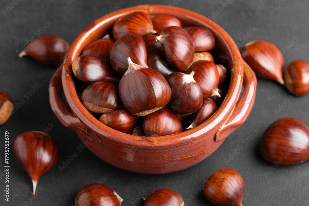 Obraz premium Raw chestnuts into a rustic clay bowl on black backdrop in warm kitchen light