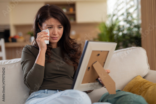 Fotografie Emotional young woman wiping tears while looking at a framed photo, expressing sadness, longing and emotional pain