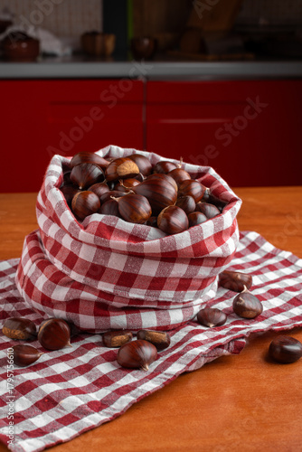 Fresh chestnuts displayed in a red checkered cloth bag on a wooden kitchen table, with scattered chestnuts creating a warm and rustic seasonal atmosphere.
