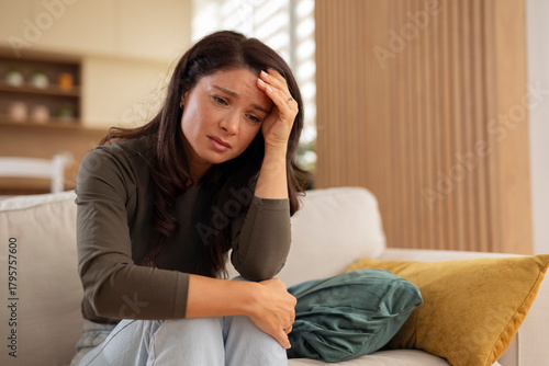 A woman sitting on a sofa, holding her head with a sad and worried expression, as if dealing with heartbreak, financial stress, or other personal problems at home.  
