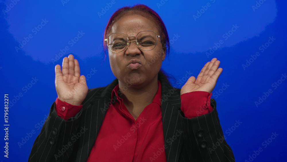 Naklejka premium African american woman covers eyes with hands while wearing glasses and red blouse in a blue studio; shyness.