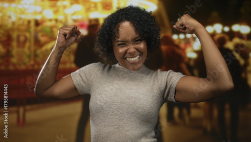 Obraz na płótnie Woman pumps fists in celebration and flashes a wide smile at a bustling street fair lined with glowing rides and stands; joy