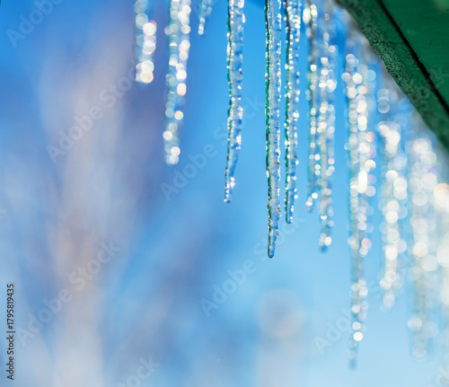A row of shiny cold icicles hang on a green roof on a sunny spring day agains...