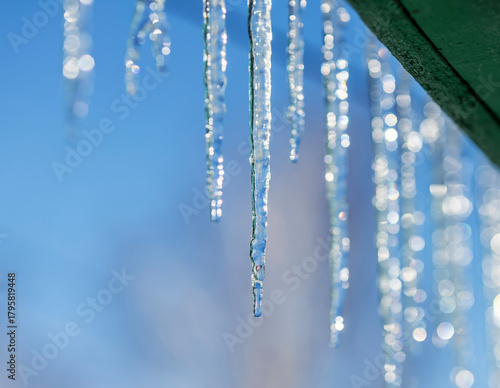 A row of shiny cold icicles hang on a green roof on a sunny spring day agains...