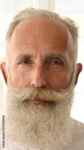 Extreme close-up portrait of a happy senior man with a large white beard. He turns, smiles at the camera, and playfully twirls his mustache. Vertical video.