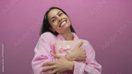 Slika na platnu Hispanic woman joyfully holding a pink gift box while wearing a robe against a vibrant pink background wall, radiating happiness and excitement