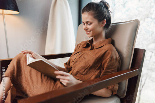 Tableau sur toile Young woman reading a book in a cozy chair, wearing a brown shirt dress, exuding