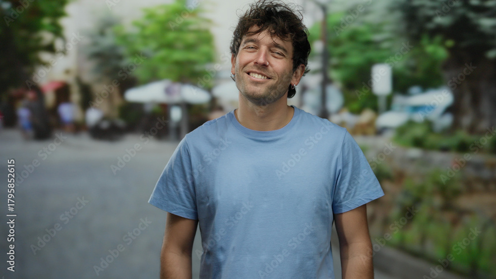 Fototapeta premium Smiling man in blue shirt stands outdoors on sunny urban street with trees and blurred buildings, exuding friendliness and confidence.