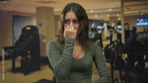 Woman pinching nose with hand and looking worried while standing in gym building near dumbbells and cardio machines; disgust unease.