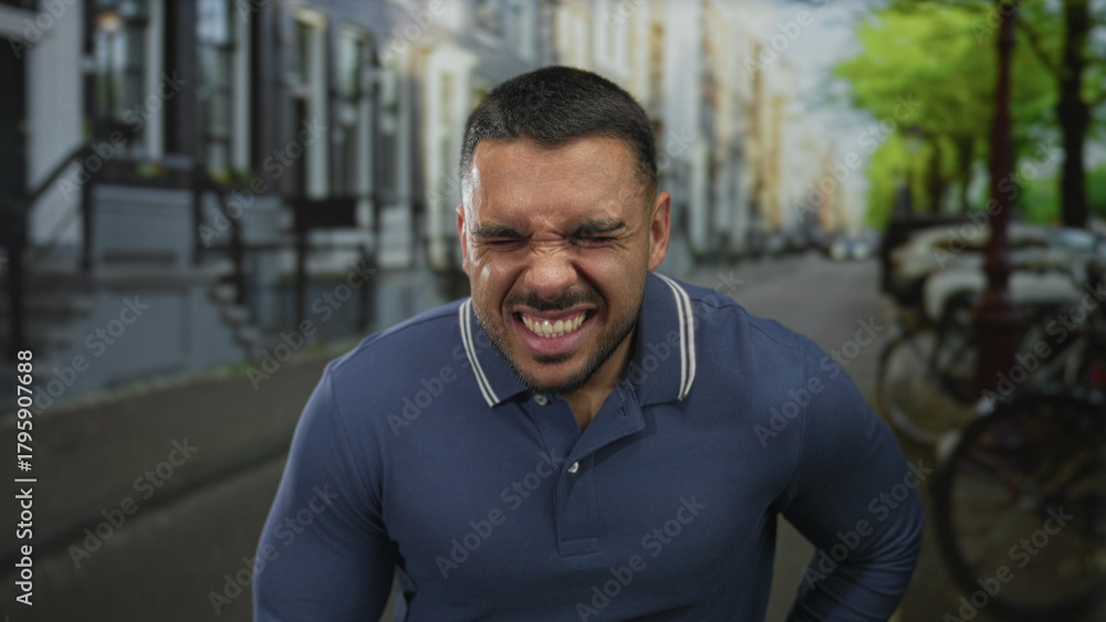 Naklejka premium Young hispanic man wearing a blue polo shirt gripping stomach and wincing on a crowded city street in daylight; pain.