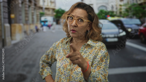 Woman points finger to camera on busy city street with parked cars and ornate building facades; blame.