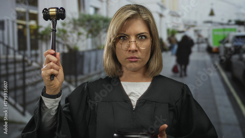 Serious woman judge in black robe holds wooden gavel aloft on bustling city street with stern expression; justice.