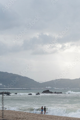 Zwei Personen stehen am Rand eines sandigen Strandes vor einer rauen See, im Hintergrund sind Hügel, Industrieanlagen und ein bewölkter Himmel mit diffusem Licht zu sehen. Die Atmosphäre wirkt ruhig u