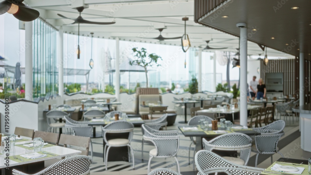 Obraz premium Soft blurred cafe interior showing a bright terrace dining area with woven chairs, tiled floor, tables, ceiling fans and glass walls; background copyspace overlay calm.