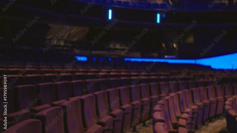Obraz premium Blurred theater seating captured in shallow bokeh interior with curved rows, dim stage glow and soft purple seats suggesting an empty auditorium; backdrop copyspace overlay template.