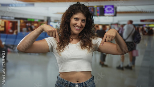 Young woman points both fingers to bare abdomen in busy airport terminal while smiling and wearing white crop top; travel confidence.