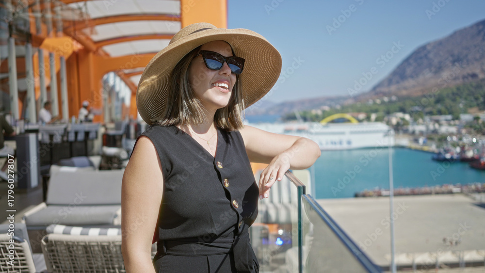Obraz premium Young woman wearing sun hat and sunglasses leaning on glass railing on building terrace smiling toward sea; relaxation vacation.