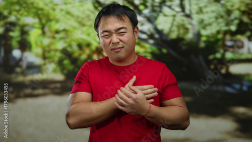 Young man in red shirt outdoors holding chest in pain against lush park background expressing discomfort and worry
