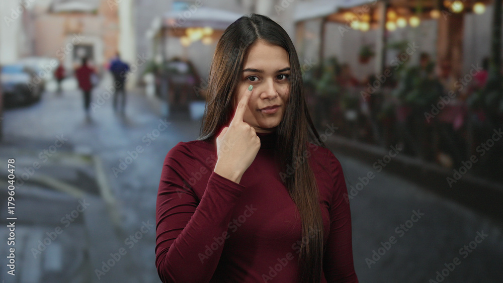 Fototapeta premium Woman pointing to her eye on a bustling terrace with blurred street background showcasing lively outdoor atmosphere in vibrant city setting.