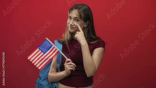 Cuadro en lienzo Young woman holding usa flag pointing finger to cheek in red studio; patriotism pride student youth