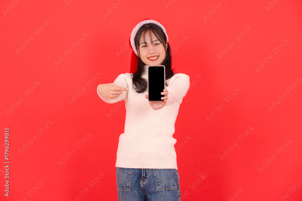 Obraz premium Asian young beautiful woman smile wearing white sweater santa hat holding show mobile smart phone blank screen on isolated on red background Merry christmas happy new year holiday festival celebration