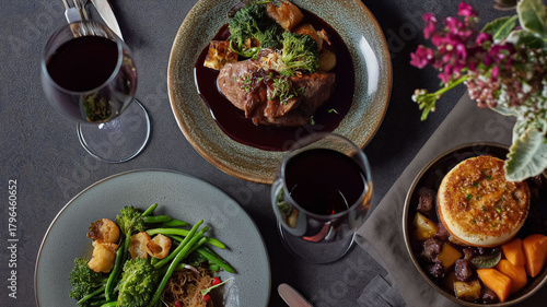 Gourmet fine dining dinner set with main course, side dish, artisan pie, and two red wine glasses on a dark table